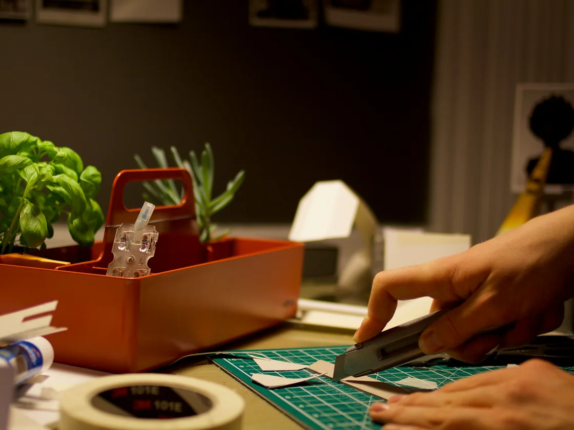 Manos sujetando un cutter sobre una base de corte verde, con recortes de papel y herramientas ordenadas en la mesa de trabajo